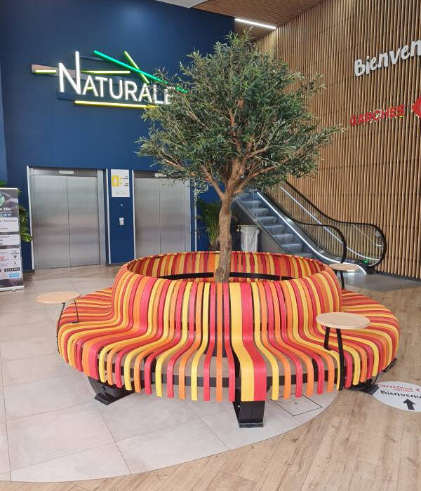 Banquette circulaire GREENWOOD en bois sur mesure avec lattes colorées et arbre central dans espace d’accueil de centre commercial