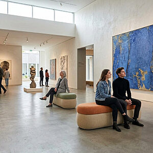 Visiteurs assis sur des bancs rembourrés SOLARA dans une salle de musée devant une grande toile bleue, mobilier d’assise pour lieux culturels